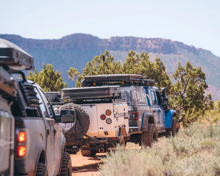 A convoy of overland vehicles towing off-road trailers through sagebrush below a mesa