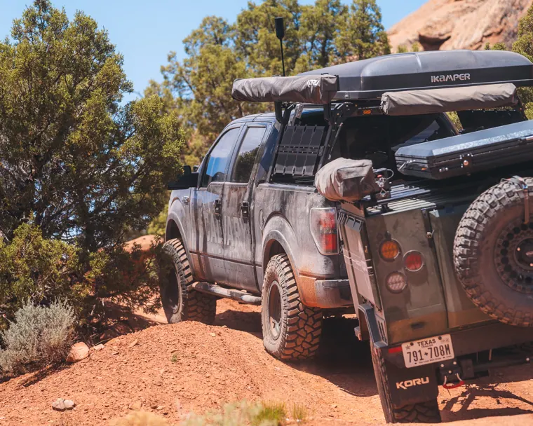 A pickup truck with a rooftop tent towing an overland trailer up a red dirt trail